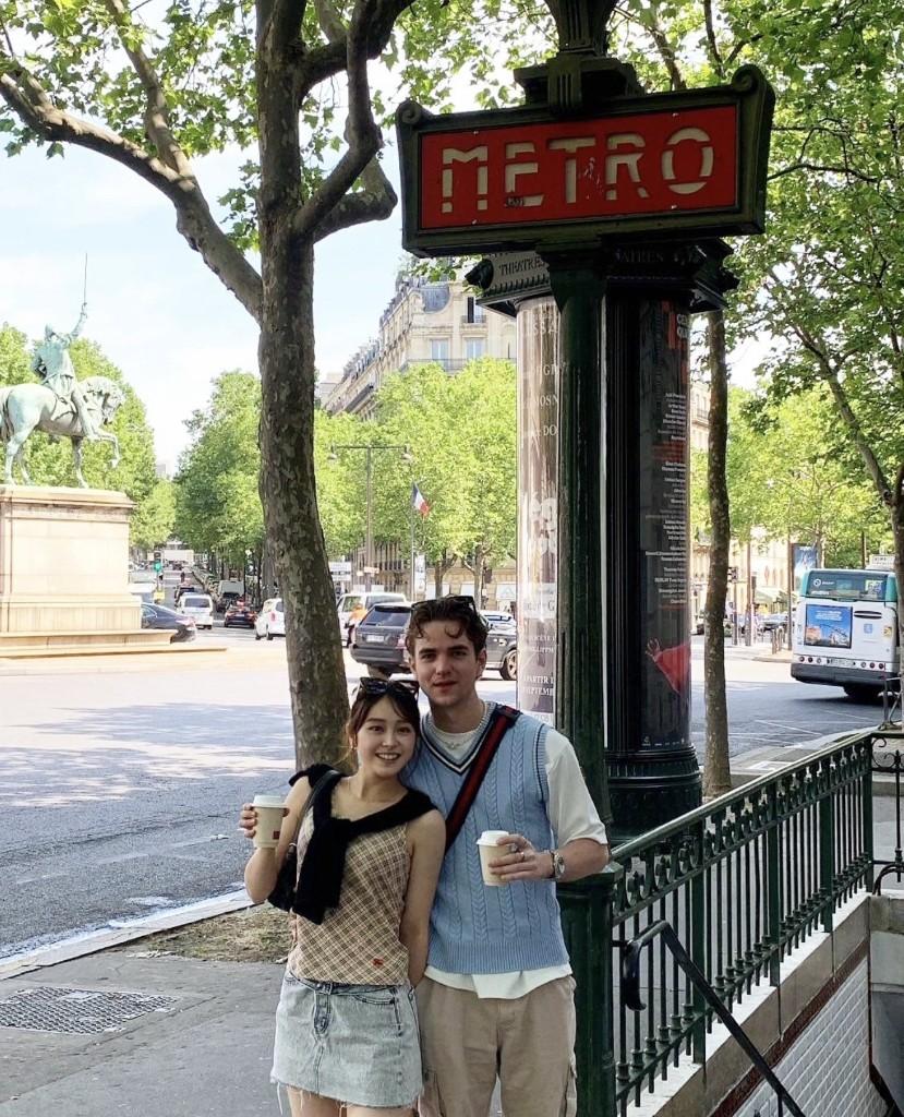 Ai and Austin with coffee cups beside a Paris Métro sign on a sunny day.
